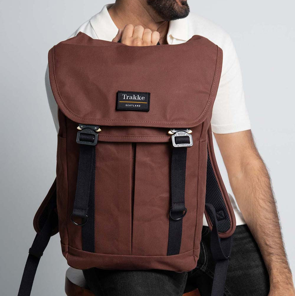 A man is sitting on a stool with the Trakke Bannoch Backpack resting on one leg. The backpack is the Port colourway and has the AustriAlpin COBRA clips.