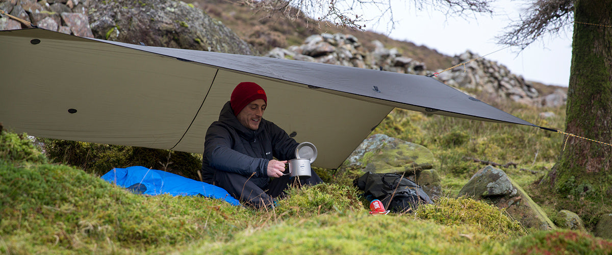 Bivvying with a camping mug under a tarp shelter on a hillside