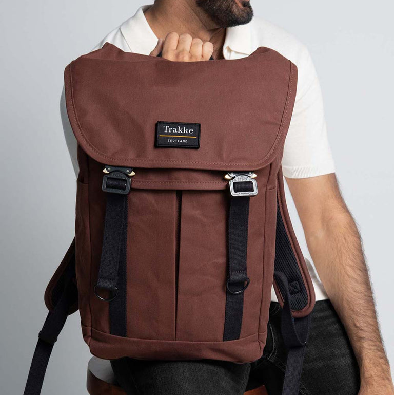 A man is sitting on a stool with the Trakke Bannoch Backpack resting on one leg. The backpack is the Port colourway and has the AustriAlpin COBRA clips.
