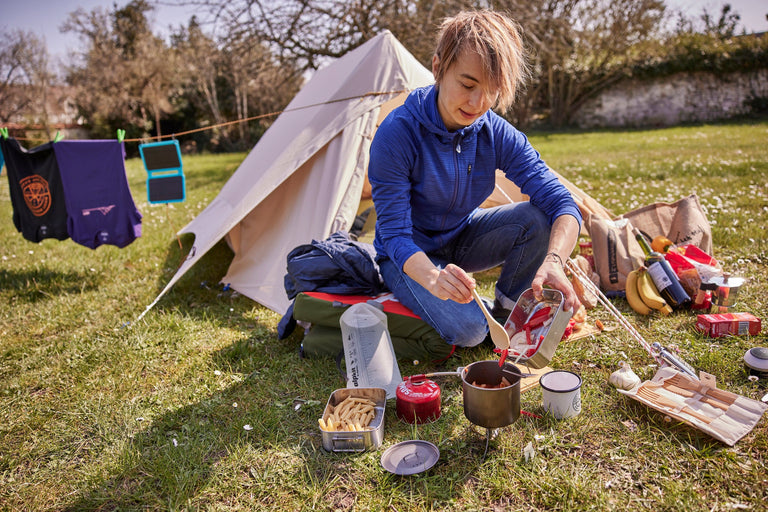 Zof sitting on a bouldering mat to cook in front of a cotton canvas tent - action