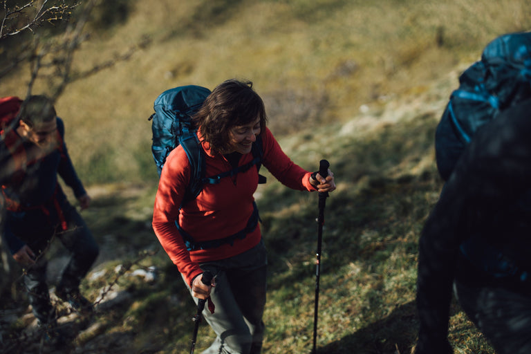 A woman hiking up a steep bank with trekking poles wearing a red fleece - action